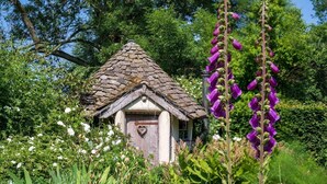 A small, rustic cottage with a thatched roof, surrounded by lush greenery and purple flowers. - The Coach House is a retreat from the outside world. (Tenbury Wells)