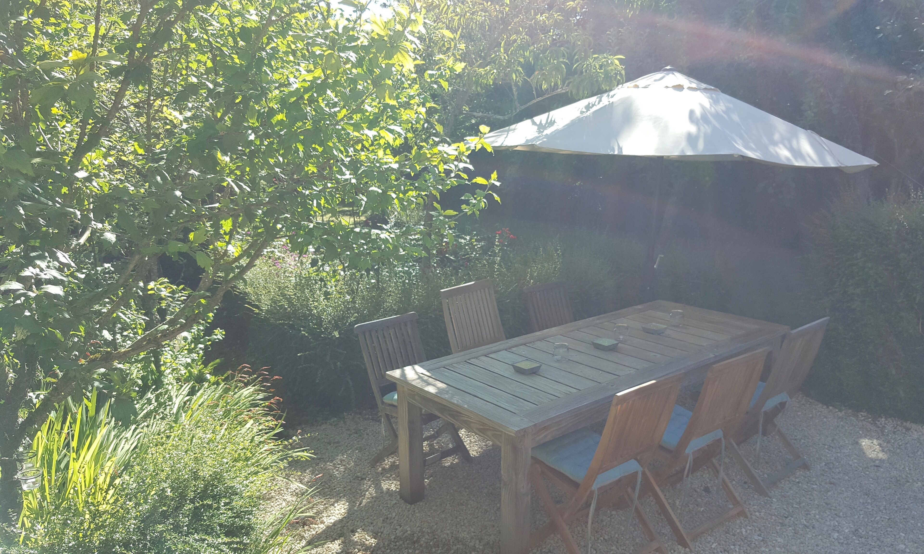 Une terrasse extérieure avec une table et des chaises en bois, entourée d’une végétation luxuriante et protégée du soleil par un parasol blanc.