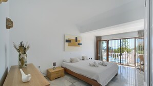 A modern bedroom with a large bed, a wooden nightstand, a vase with dried flowers, and a view of a balcony with a pool.