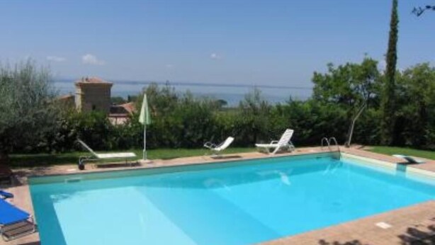 Una piscina rectangular con agua azul cristalina, rodeada por una terraza empavedada. Hay dos sillas de descanso y una sombrilla en la terraza.