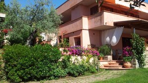 Un edificio de dos pisos con un jardín delantero, que incluye un camino pavimentado y diversas plantas y flores.