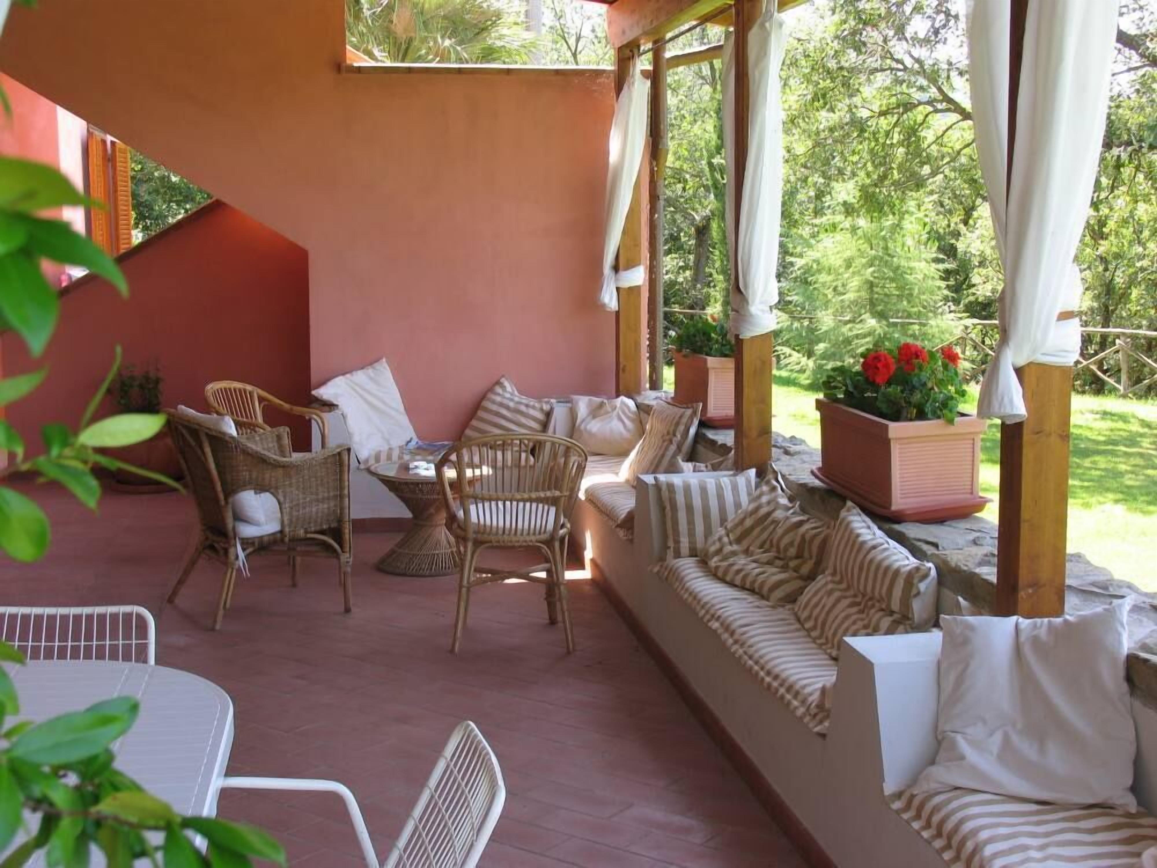 A covered outdoor seating area with wicker furniture and a cushioned sofa, surrounded by greenery.