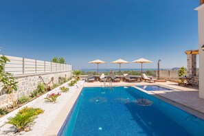 A pool with lounge chairs and umbrellas, a stone wall, and a view of the sea.