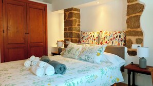 Une chambre à coucher avec un lit, une armoire en bois, des murs en pierre, une couvre-lit à motifs floraux et des serviettes.