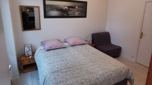 Un dormitorio con cama, una silla, una mesita de noche y un espejo, además de un cuadro colgado en la pared.