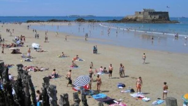 Una playa con gente tomando el sol y nadando, una estructura que parece un castillo al fondo, y un cielo despejado.