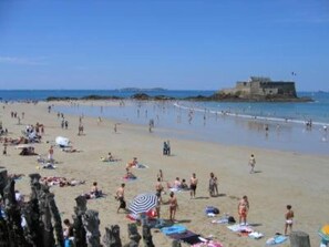 Una playa con gente tomando el sol y nadando, una estructura que parece un castillo al fondo, y un cielo despejado.