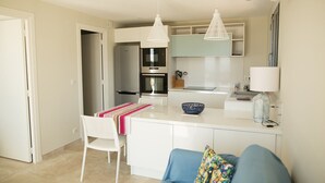 A modern kitchen with white cabinetry, a built-in oven, and a microwave. There is a dining area with a white table and chairs, and a blue sofa with colorful cushions. - Villa sea view heated pool 8 persons with electric charging point (Sainte Lucie de Porto Vecchio)