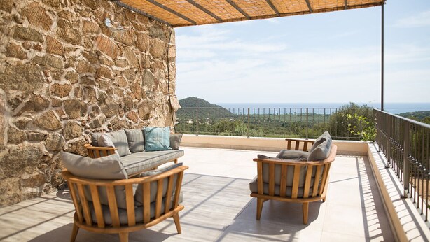 A balcony with stone walls, two wooden chairs, a sofa, and a view of the sea. - Villa sea view heated pool 8 persons with electric charging point (Sainte Lucie de Porto Vecchio)