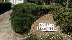 A sign reading "CASINHA LECOTEC" in a garden area with a gravel path, bushes, and a building in the background.