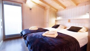 Una habitación con dos camas, cada una con una manta oscura y una toalla doblada. La habitación tiene paredes de madera y un ventanal grande.