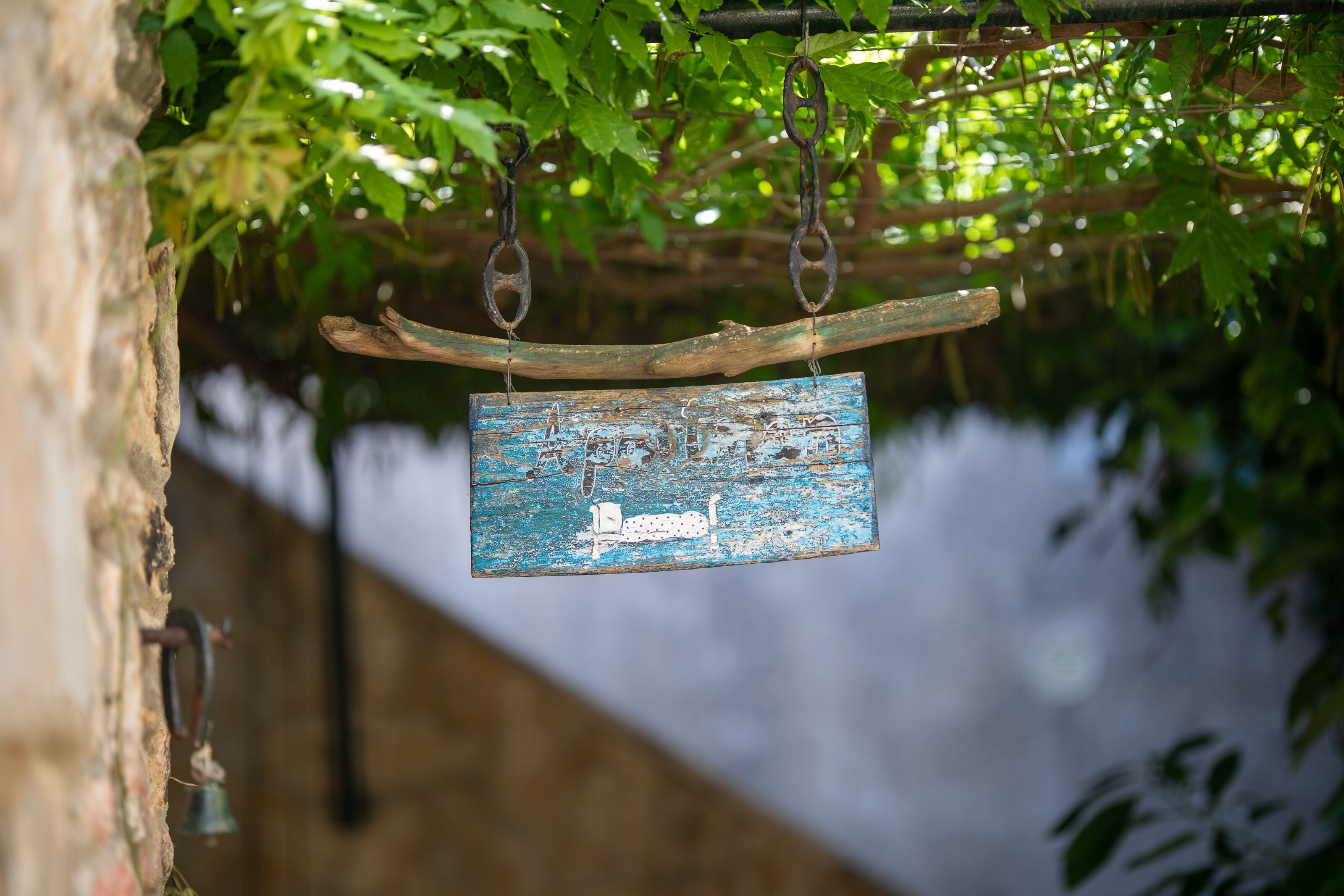 Ein rustikales Holzschild mit einer verblichenen Illustration, das an einem Metallhaken befestigt und an einer Steinwand mit einer Glocke angebracht ist.