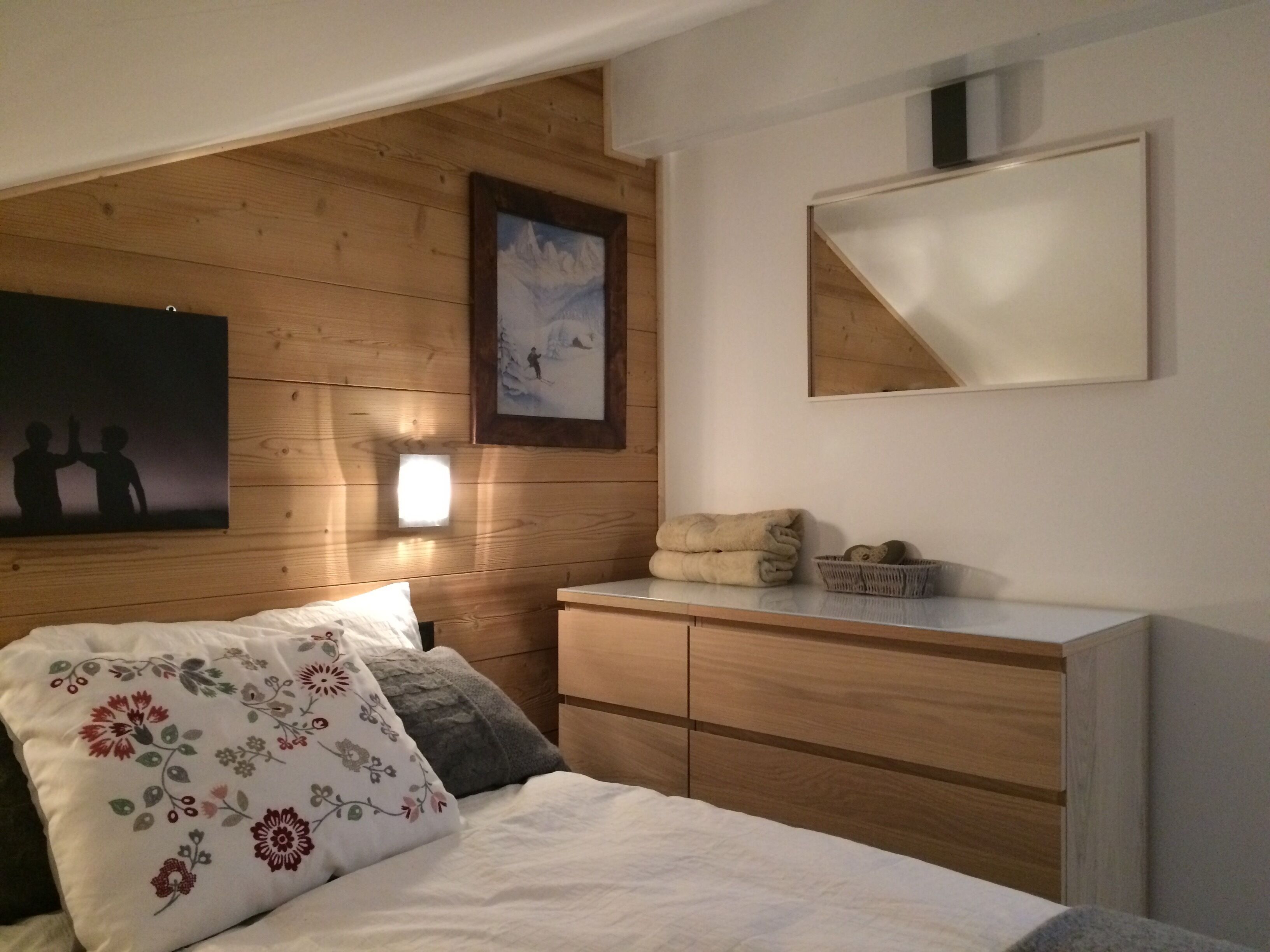 A bedroom with a wooden headboard, a bed with floral pillows, a nightstand, a dresser, and a framed picture on the wall.