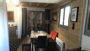 A dining area with a wooden table, chairs, and a red cushion. There is a kitchen in the background with cabinets and a refrigerator. - Apartment Harikoa, sleeps 5, next to piste, Col de Merdassier. (Manigod)