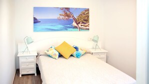 Une chambre à coucher comprenant un lit, deux tables de chevet, une commode, un tableau représentant une scène de plage et une fenêtre.