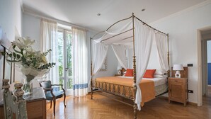 A four-poster canopy bed with a wooden frame, a nightstand with a lamp, and a chair in a room with wooden flooring and a large window with sheer curtains.