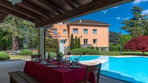 A covered outdoor dining area with a table set for a meal, overlooking a swimming pool and a building with a red roof.