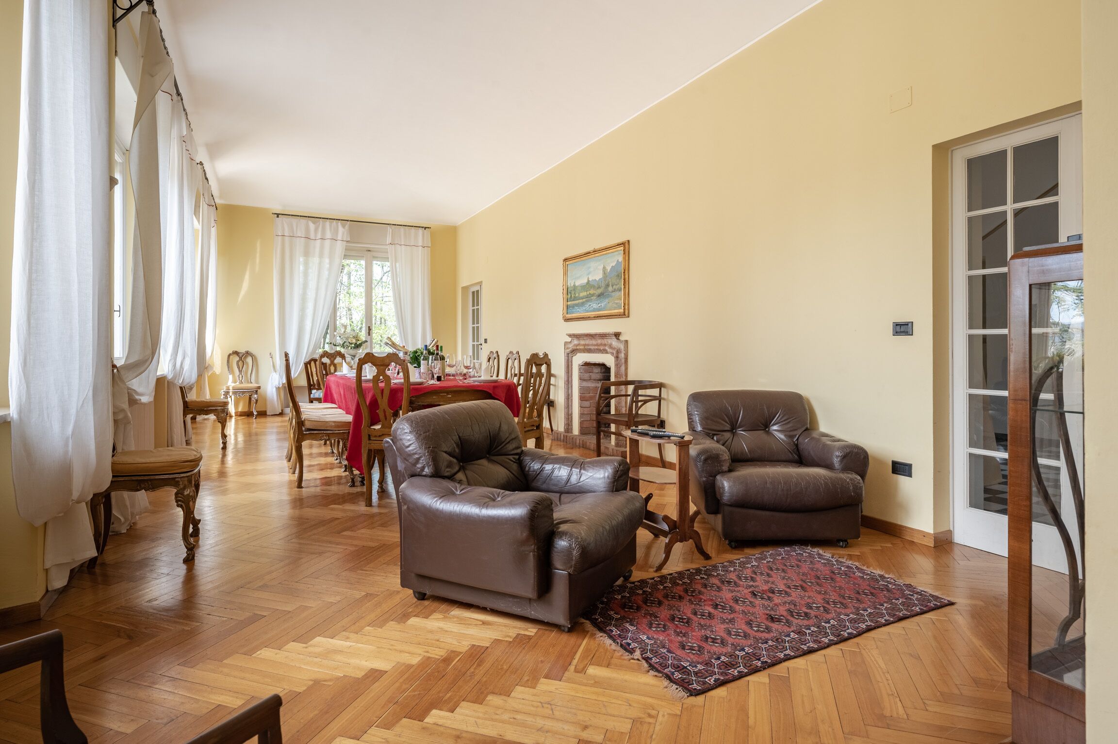 Un salon avec deux fauteuils en cuir, une table basse en bois et un tapis rouge. On aperçoit à l’arrière-plan un coin repas avec une table et des chaises rouges.