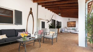 Ein geräumiger Wohnbereich mit einer Couch, zwei Sesseln, einem Couchtisch und einem Esstisch. Eine Holzbalkendecke, ein großes Fenster mit Rollläden und ein Gemälde an der Wand sind ebenfalls vorhanden.