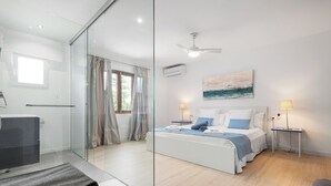 A modern bedroom with a glass partition, a bed with blue bedding, a desk with a chair, and a ceiling fan.
