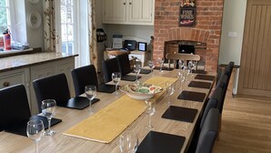 A dining area with a long wooden table, black chairs, and wine glasses. There is a brick wall with a fireplace and a clock on the wall. - Large open plan cottage in conservation village just outside York. 3 night deals (York)