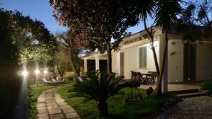A well-lit outdoor area with a paved walkway, lounge chairs, and a dining table set up under a covered patio.