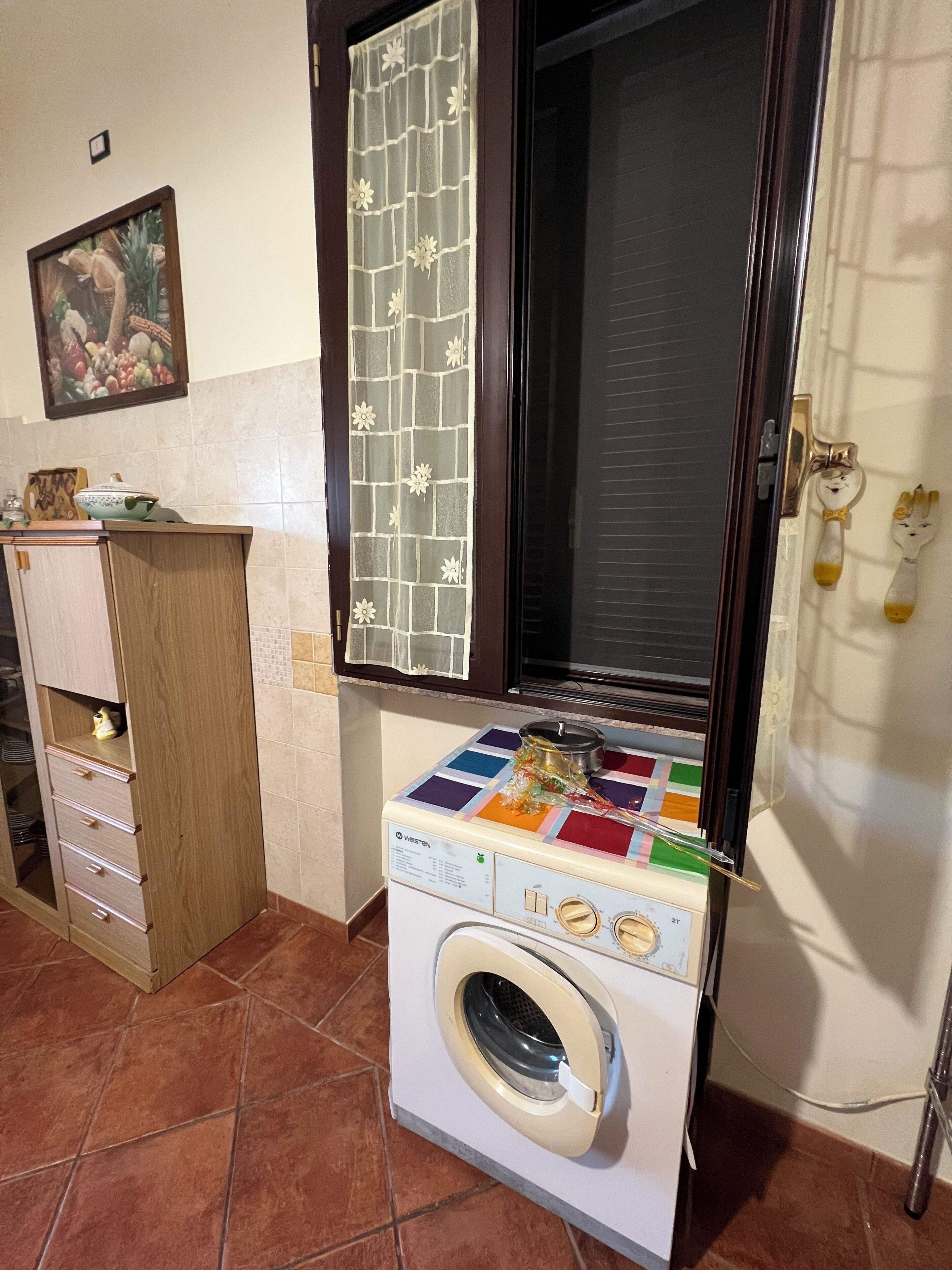 A compact kitchen with a washing machine, a cabinet with drawers, a tiled backsplash, and a window with a decorative screen.