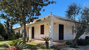 A white building with a tiled roof and black shutters, surrounded by well-maintained greenery and a stone pathway.