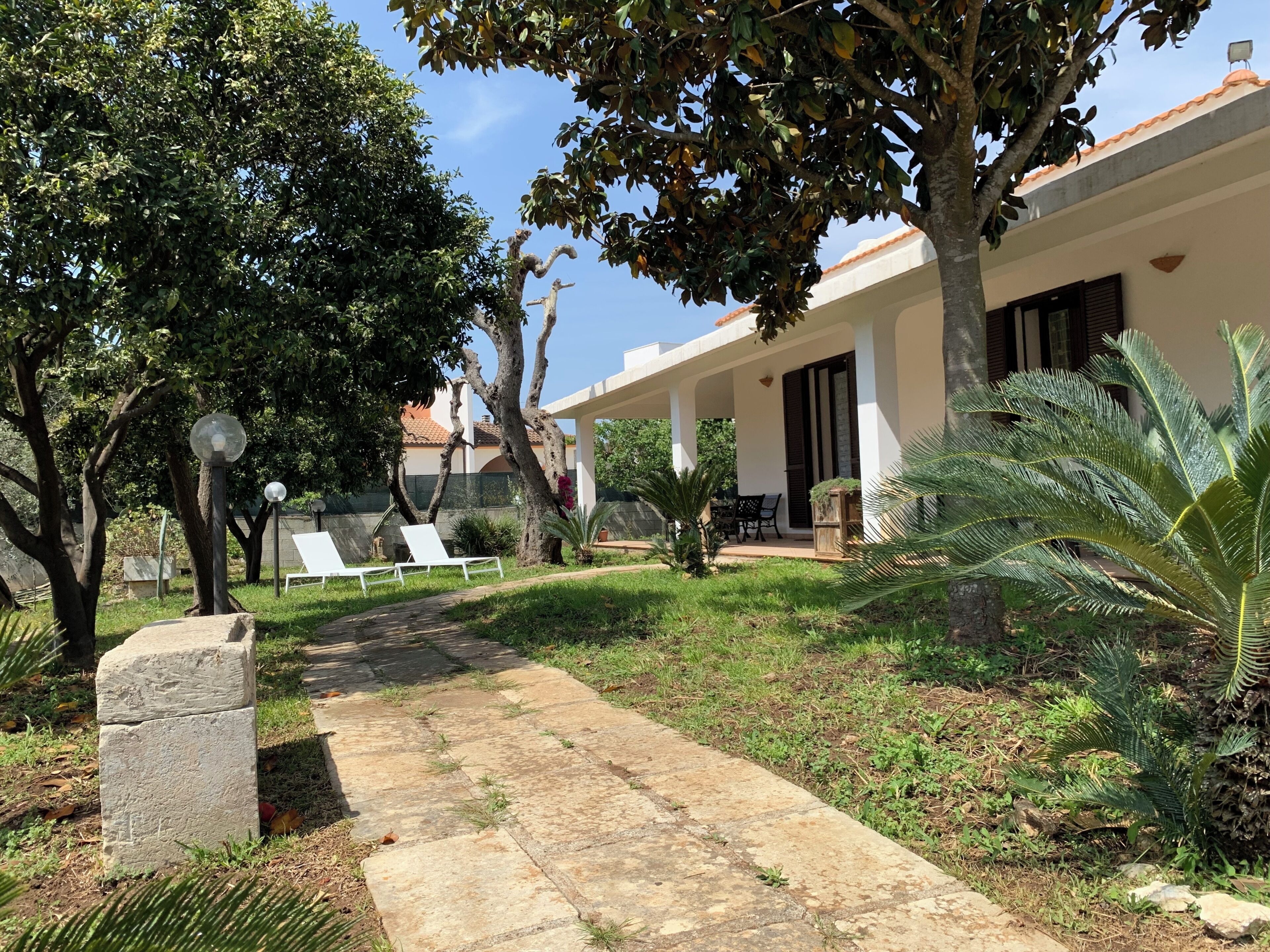 Un chemin mène à un bâtiment doté d’un patio couvert, d’un espace extérieur aménagé pour s’asseoir et d’un jardin bien entretenu où poussent différentes espèces de plantes.