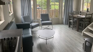 A modern living room with a sofa, two armchairs, a small coffee table, and a dining table with chairs. There are large windows with curtains, a clock on the wall, and a ceiling light fixture. - Detached holiday home in Westkapelle for 4 people. (Westkapelle)