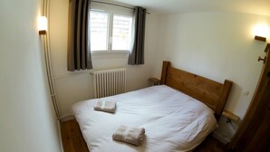 Une chambre à coucher avec un lit en bois, du linge de lit blanc, une fenêtre avec des rideaux, un radiateur et un plancher en bois.