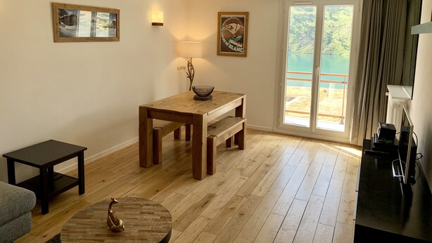 Une pièce comprenant une table à manger en bois, une petite table ronde agrémentée d’un objet décoratif, un canapé et une vue sur un lac, visible à travers une porte-fenêtre coulissante.