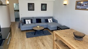 Un salon moderne avec un canapé d’angle gris, une table basse ronde, une table à manger en bois et une cuisine en arrière-plan.