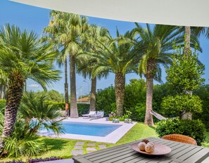 Een zwembadterras met palmbomen, een zwembad, ligbedden en een tafel met decoratieve objecten.