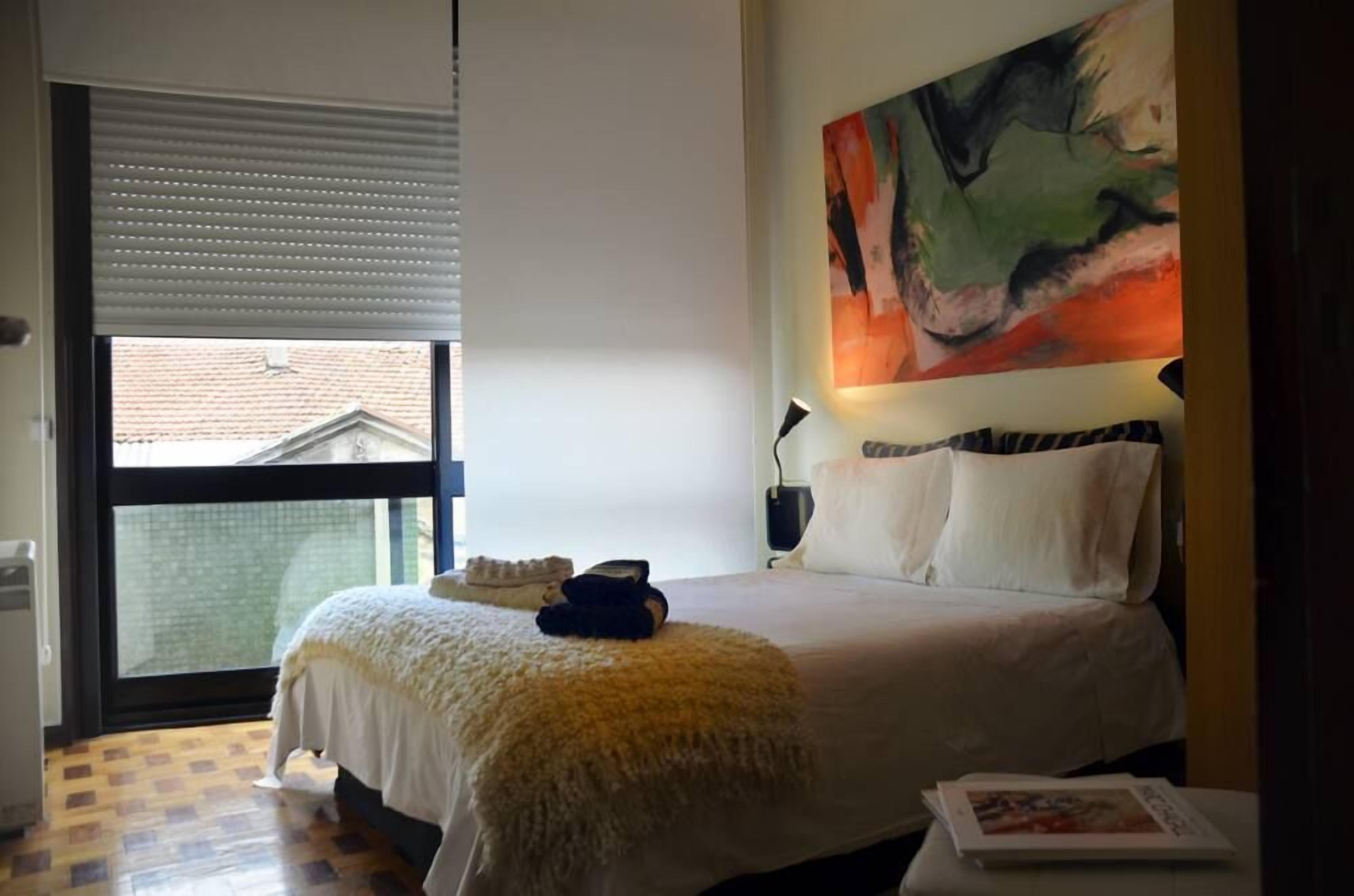 Ein Schlafzimmer mit einem großen Bett, einem Fenster mit Jalousien, einem Bild an der Wand und einem Nachttisch mit Lampe.