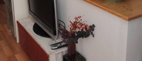 A living room with a flat-screen TV on a wooden stand, a red basket on a white cabinet, and two vases with flowers on a wooden shelf.