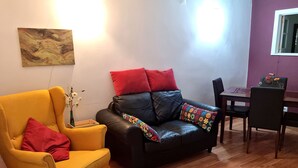 Een woonkamer met een geel fauteuil, een zwarte leren bank met rode en kleurrijke kussens, een houten salontafel en een eettafel met stoelen.