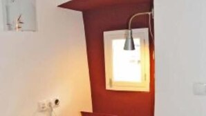 Een kleine kamer met een houten tafel, een rode stoel, een raam met lamp en een witte muur.
