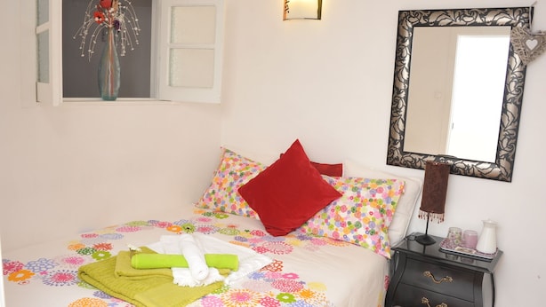 Een netjes opgemaakt bed met kleurrijke bloemenbedden, een rode kussen en een groene handdoek. Er staat een klein nachtkastje met een lamp en een spiegel met een sierlijk frame.