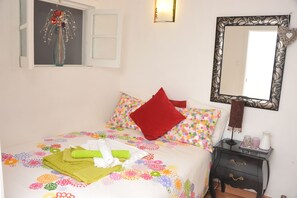 Ein ordentlich bezogenes Bett mit bunter Blumenwäsche, einem roten Kopfkissen und einem grünen Handtuch. Ein kleiner Nachttisch mit Lampe und einem Spiegel mit aufwendigem Rahmen sind ebenfalls vorhanden.