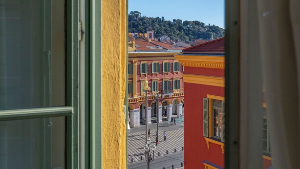 Une vue depuis une fenêtre donnant sur un mur jaune, avec en contrebas une rue bordée de bâtiments aux couleurs vives et un ciel bleu dégagé.