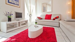 Un salon moderne avec un canapé blanc, des coussins rouges, une table basse blanche et un téléviseur posé sur un meuble blanc.