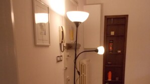 Kapea käytävä, jossa on oranssilla tyynyllä varustettu tuoli, lattiavalaisin, patteri ja hylly, jossa on erilaisia esineitä.
