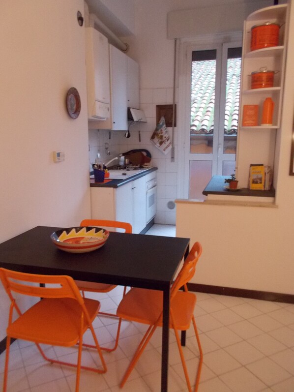 A compact kitchen with white cabinets, a small dining table with orange chairs, and a window with a view of a tiled roof.