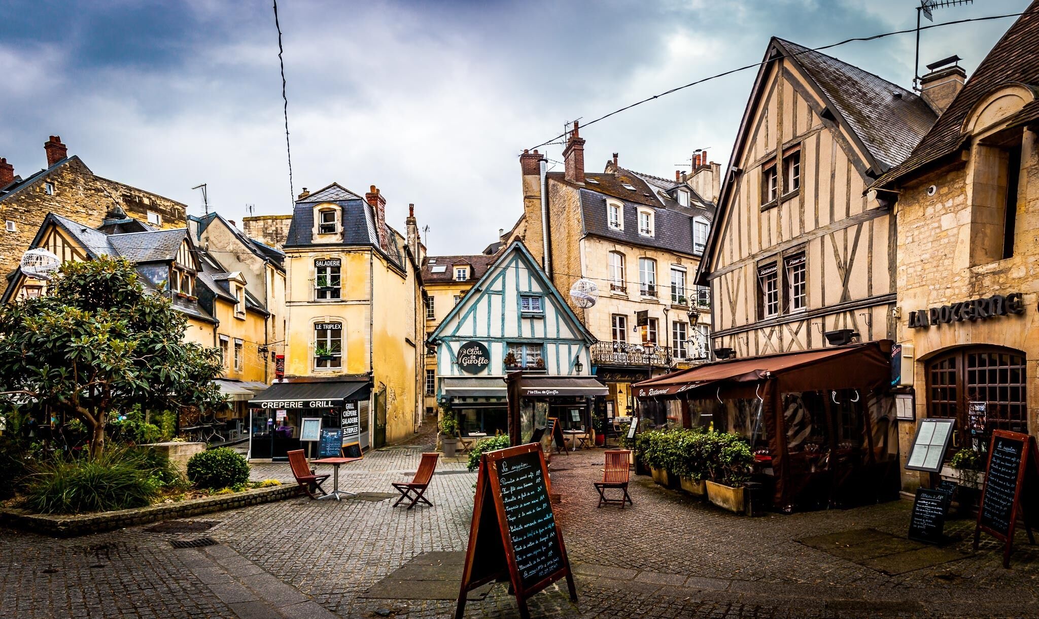 Une rue pavée bordée de bâtiments européens traditionnels, avec des tables en terrasse et une enseigne indiquant l’emplacement d’un restaurant.
