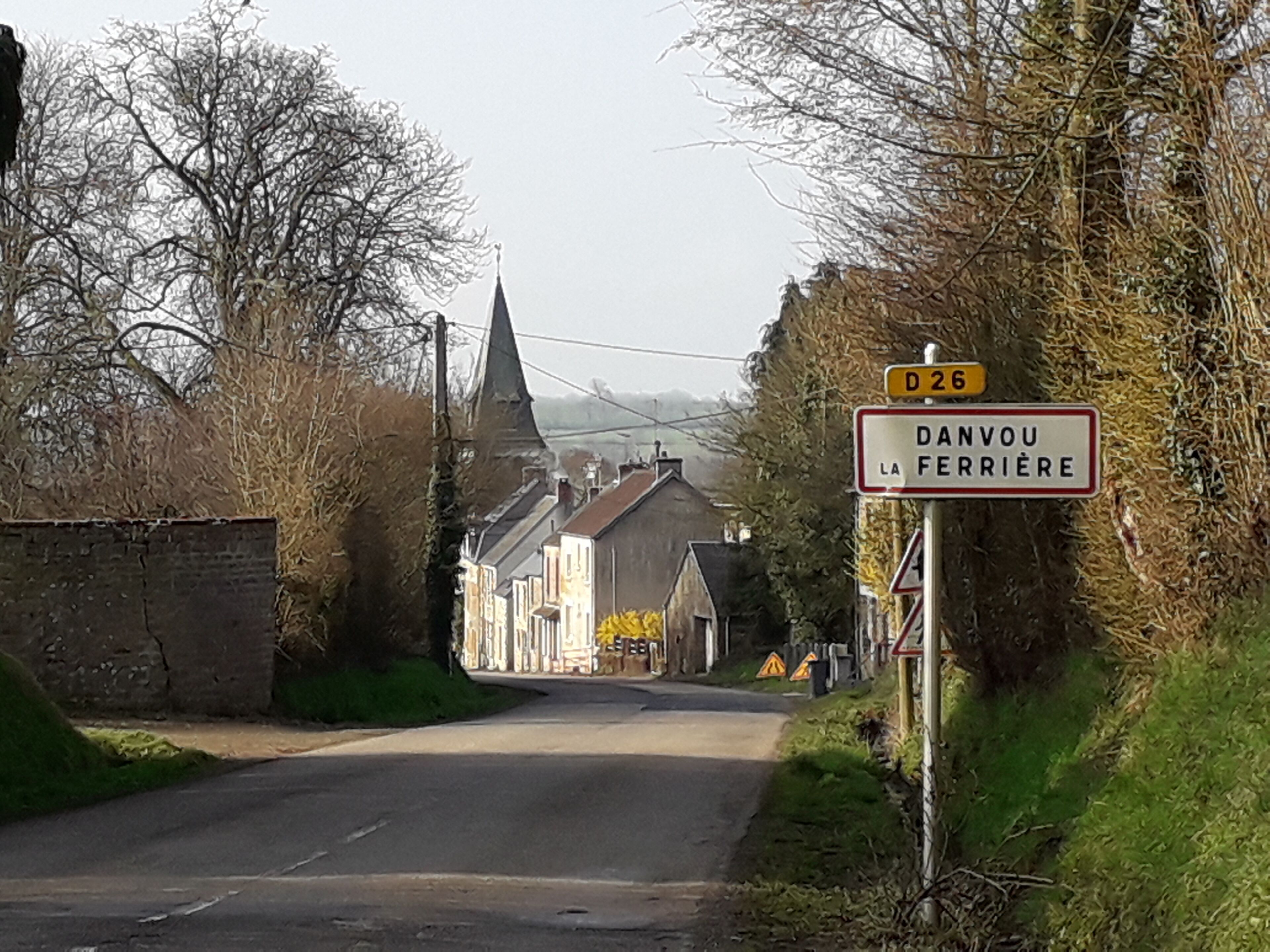 Un camino rural con la aguja de una iglesia a lo lejos, un cartel que indica 'DANVOU LA FERRIÈRE' y una señal de advertencia amarilla.