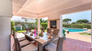 Un patio exterior con una mesa de comedor, sillas de mimbre, una piscina y una pantalla de televisión.