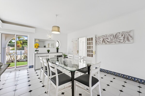 A modern dining area with a glass table, white chairs, and a view of a garden through glass doors. - Villa Jade | 4 Bedrooms | Pool | Old Village | Vilamoura (Vilamoura)