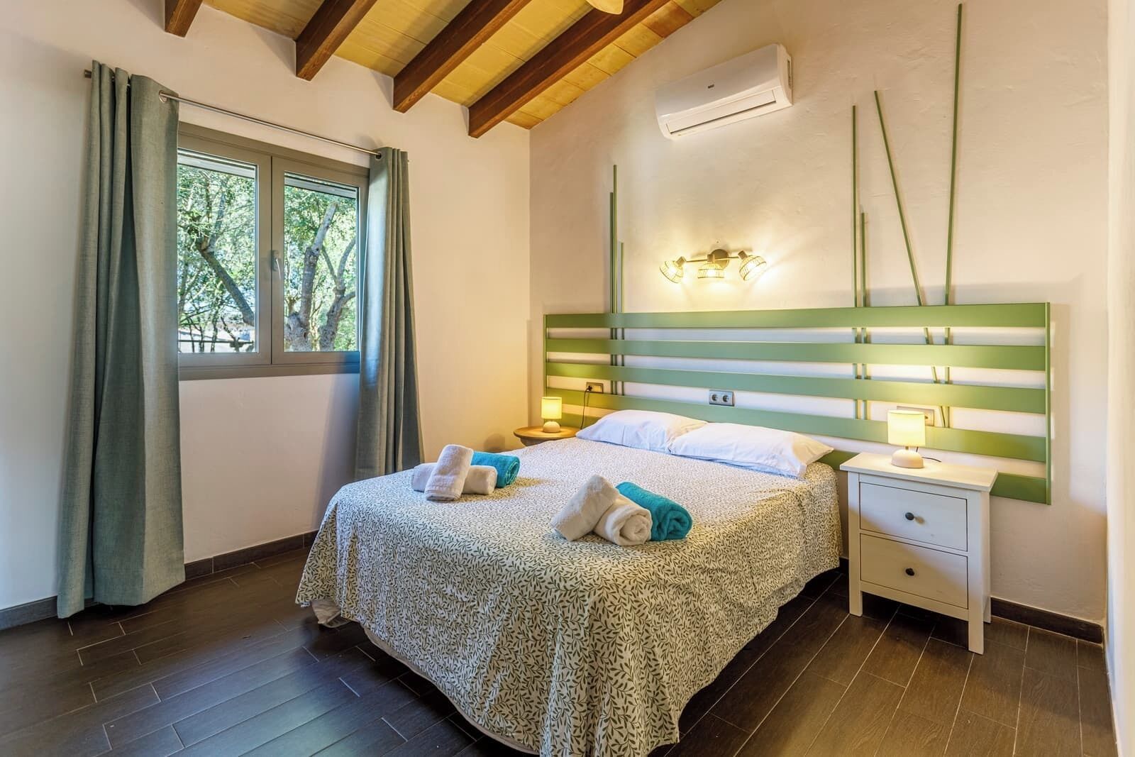 Un dormitorio con cama, mesitas de noche, aire acondicionado y ventana con cortinas.