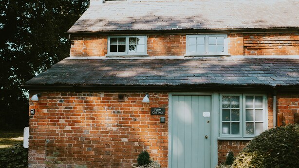 Een bakstenen huisje met een groene deur, een klein raam en een bordje met de tekst 'PLUM COTTAGE'.